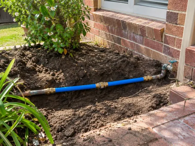 Exposed blue repaired water pipe in a garden trench beside a brick house wall in Lindfield